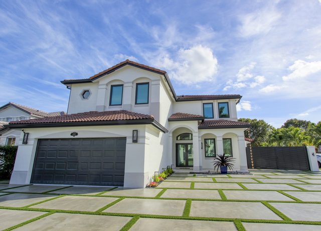 White two-story home with a barrel tile roof, seen from a driveway of concrete slabs set in strips of grass, under a blue sky.