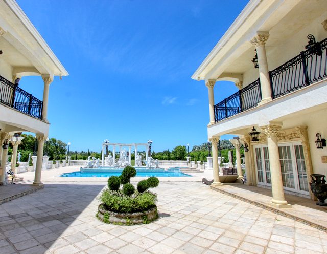 Wide courtyard of a columned estate, with a swimming pool and a colonnade of statues at the far end.