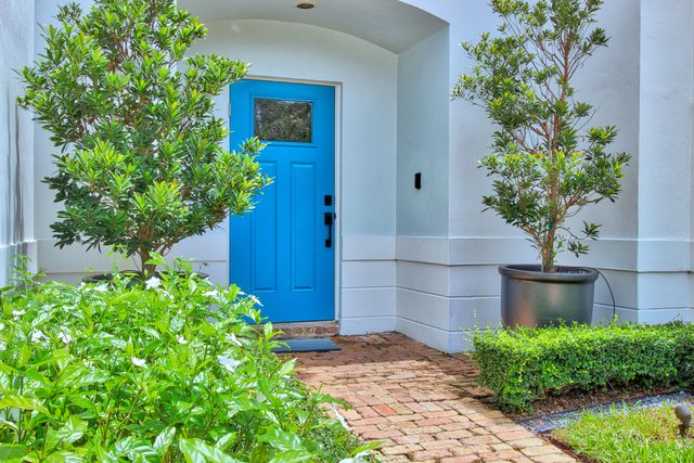 Bright blue front door under a curved porch, with white gardenias in flower beside a brick path.