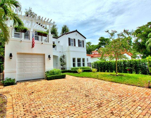 White two-story home with shuttered windows and a covered garage, seen across a herringbone brick driveway.