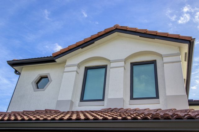 Gable end of a house seen from below, with two dark-framed arched windows and a barrel tile roof against a blue sky.