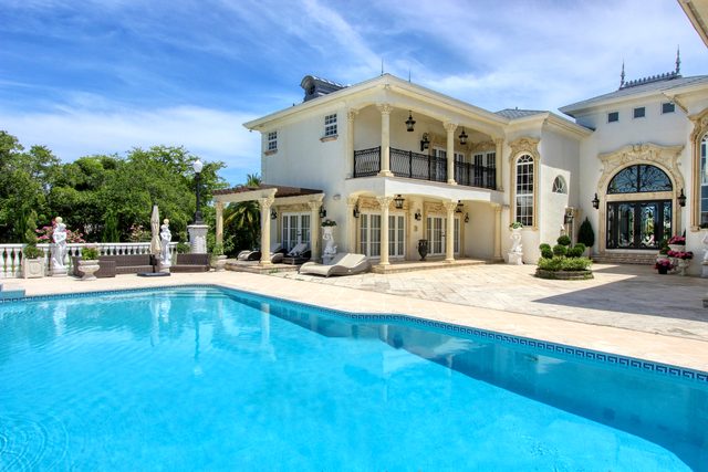 Long swimming pool running along the rear elevation of a columned estate, with balconies above the arched openings.
