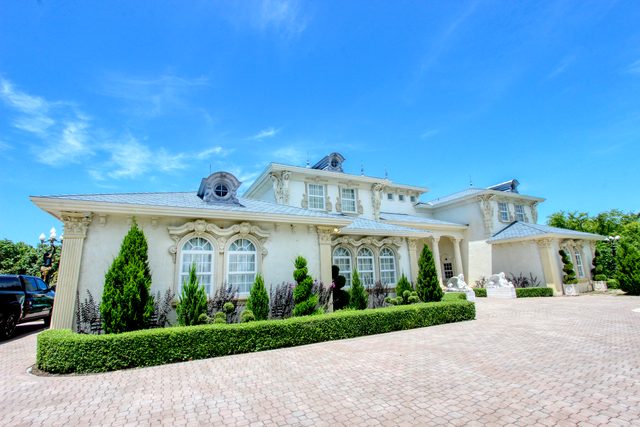 Wide front elevation of a columned estate under a deep blue sky, with clipped hedges along a paver driveway.