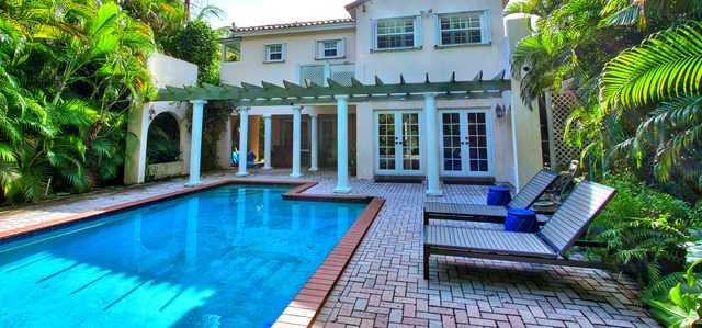Swimming pool edged in brick paving, with a pergola along the house and lush palms behind.