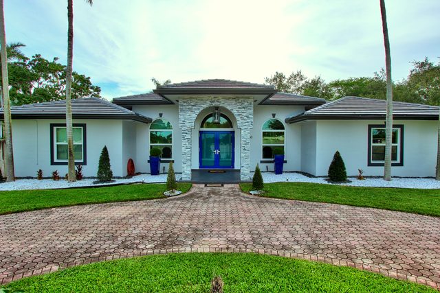 Single-story white house with blue double doors in an arched stone-clad entry, framed by palms.