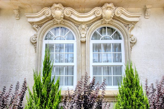Pair of tall arched windows framed by carved stone surrounds, with clipped cypress trees below.